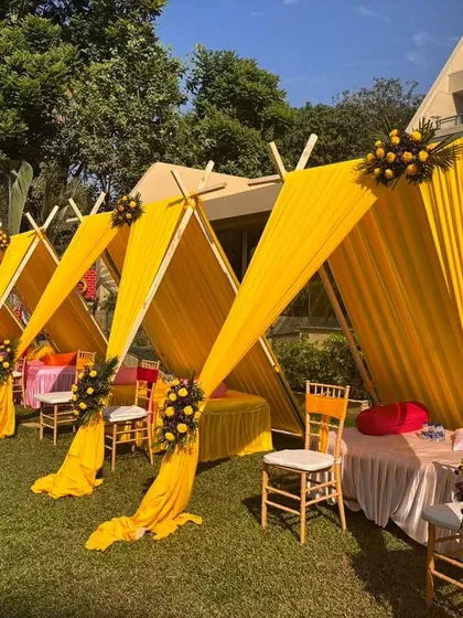 Bright yellow draped cabanas provide stylish and comfortable seating for guests at this outdoor Haldi function. It's a practical and aesthetic solution for a sunny day.