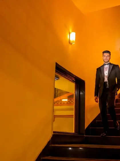 The groom descends a staircase, captured in a full-length shot with warm, dramatic lighting.