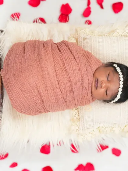 An overhead view of a newborn sleeping on a miniature bed, surrounded by scattered red rose petals. The simple elegance of this shot makes it a beautiful keepsake.