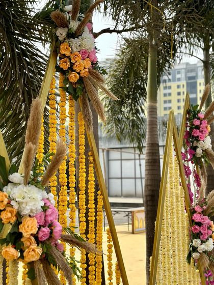 A perfect Haldi decor setup with triangular structures adorned with marigold strings and bouquets of roses and pampas grass, blending traditional and modern aesthetics.