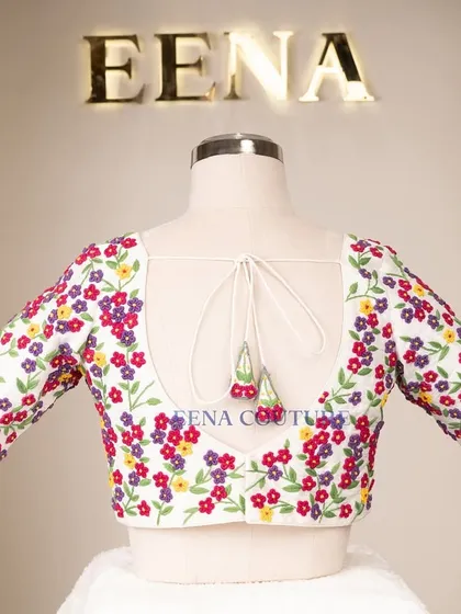 A garden of flowers on a blouse. This white raw silk piece is covered in vibrant, multi-colored floral French knot embroidery, making it a cheerful and eye-catching addition to any festive wardrobe.