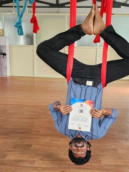 An aerial yoga student joyfully displays his certificate while hanging upside down, capturing the fun spirit of the practice.