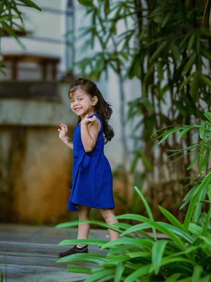 A candid shot of the little girl in the blue dress, looking back with a sweet smile. The lush green foliage frames her beautifully in this natural portrait.