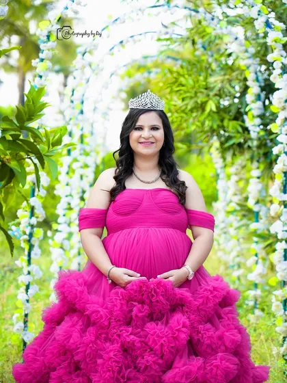 A "Barbie" themed portrait of a mother-to-be in a vibrant pink ruffled gown and crown, under a floral arch.