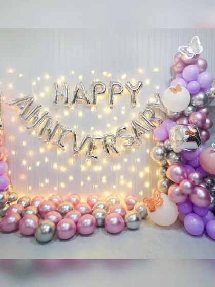 A "Happy Anniversary" decoration with a fairy light wall. The purple and silver chrome balloons add a touch of glamour to this romantic home setup.