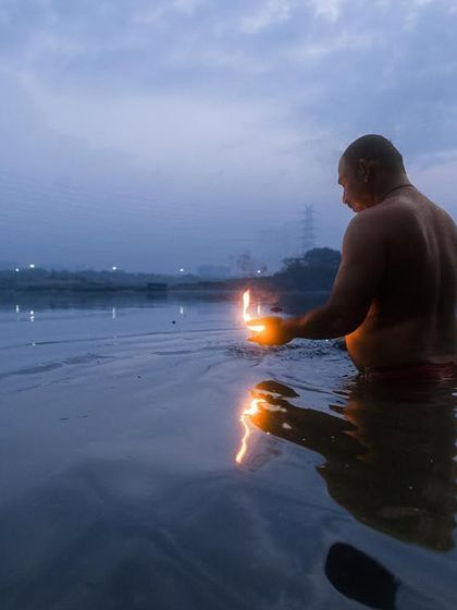 A man performs a ritual with a lit diya in the Yamuna river at dusk, his reflection shimmering on the water's surface.