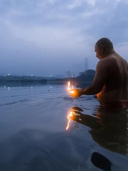 A man performs a ritual with a lit diya in the Yamuna river at dusk, his reflection shimmering on the water's surface.