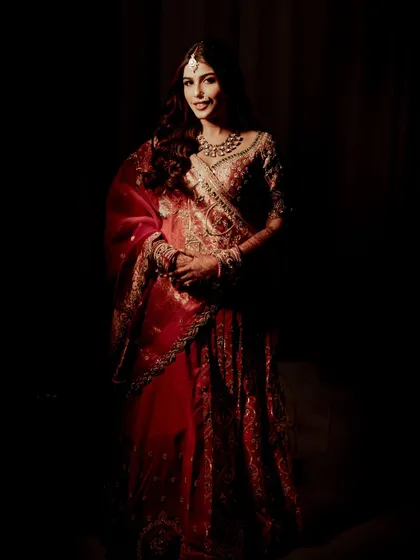 A full-length portrait against a dark background, emphasizing the grandeur of the red bridal lehenga. The lighting focuses solely on the bride, making her the center of this artistic shot.