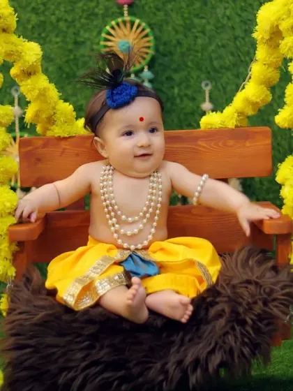 A happy baby dressed as Krishna, sitting confidently on the swing. This shot showcases the full costume, including the peacock feather headband and pearl jewelry.