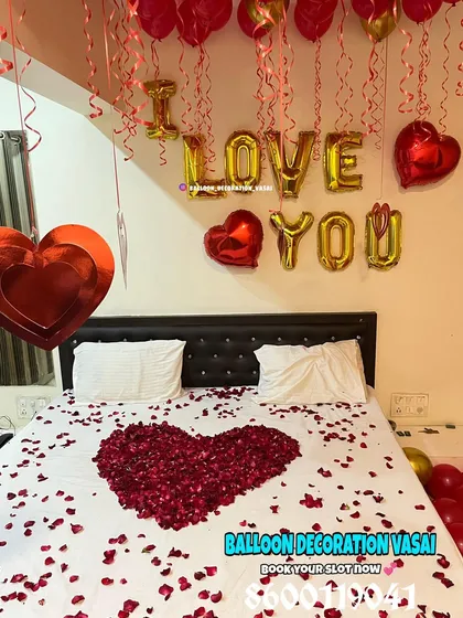 This romantic bed decoration features a heart made of red rose petals, surrounded by more petals and "I LOVE YOU" foil balloons on the headboard. Red and gold balloons float on the ceiling.