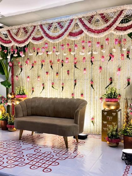 An elegant backdrop for a home ceremony, featuring a curtain of hanging jasmine and pink lotus buds. The decor is completed with banana plants and a traditional kolam on the floor.