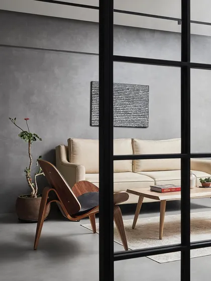 A view of the living room through a custom black-framed glass partition. The iconic shell chair adds a classic mid-century touch, complementing the custom ash wood coffee table and the minimalist aesthetic of the space.
