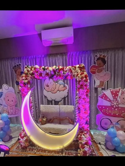 A home baby shower decoration with a floral swing and lighted moon. The setup is flanked by 'It's a Boy' and 'It's a Girl' props.