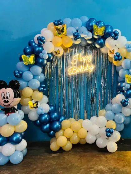 A Mickey Mouse themed first birthday with a circular balloon arch against a blue wall. The setup is accented with a neon sign, a Mickey balloon column, and a light-up number '1'.