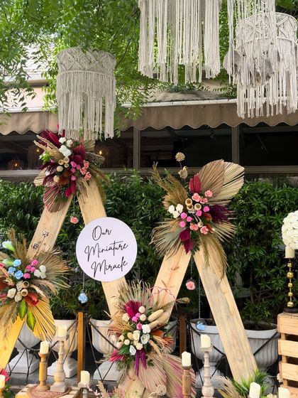 A unique and artistic backdrop for a baby shower. This boho-chic design uses triangular wooden frames adorned with dried palm leaves and vibrant floral bouquets, with a "Our Miniature Miracle" sign.