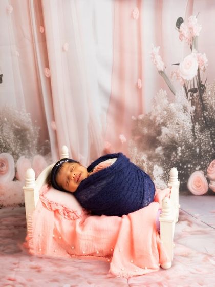 A smiling newborn sleeps in a miniature white bed, set against a beautiful floral pink backdrop for a dreamy, romantic feel.