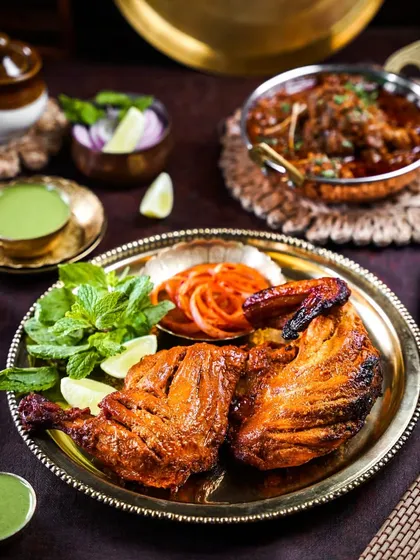 A classic Tandoori Chicken served on a brass plate with mint chutney and onion salad. The rustic, dark styling highlights the charred texture and vibrant red color of the dish.