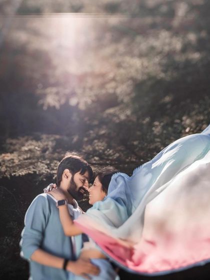 An artistic shot with the bride's colorful saree flowing in the wind. The motion blur and sun flare add a dreamy, ethereal quality to this romantic embrace in the woods.