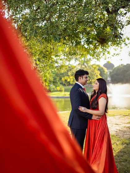 A romantic embrace framed by lush greenery and the flowing trail of the orange-red gown. This is a perfect example of a classic, beautiful pre-wedding portrait.