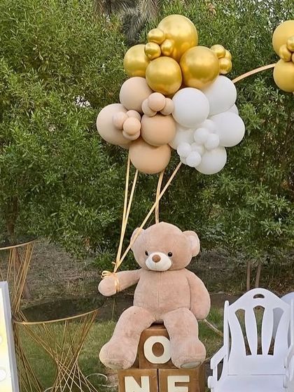 A close-up of the teddy bear detail from a first birthday party. The teddy bear is seated on 'ONE' blocks and appears to be holding a bunch of gold, beige, and white balloons.