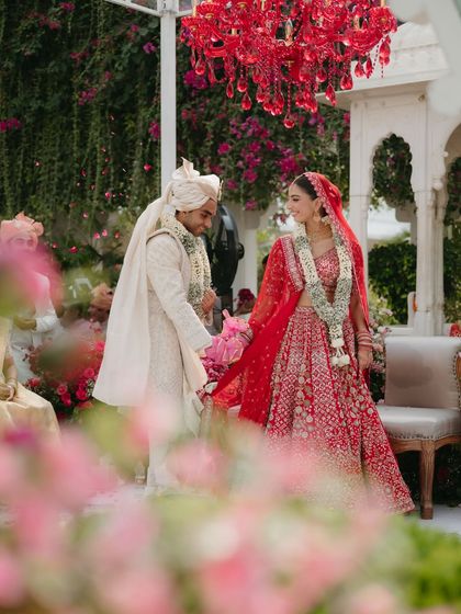 A candid moment between the couple during their pheras. The frame is filled with soft florals and the striking red chandelier, capturing the romance and grandeur of their special day at Taj Lake Palace.