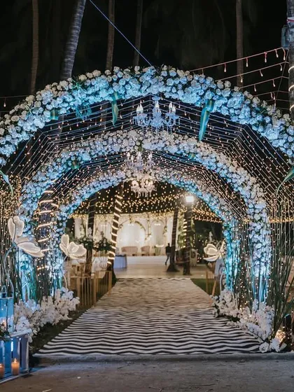 A magical "under the sea" or "winter wonderland" themed entrance tunnel. The arch is covered in white flowers and illuminated with blue lights and fairy light curtains for a truly immersive experience.