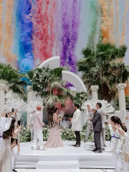 A moment of pure, unadulterated joy. For this elegant white wedding, a burst of rainbow-colored smoke created a stunning, unexpected backdrop, proving that even the most serene celebrations can have a vibrant, playful heart.