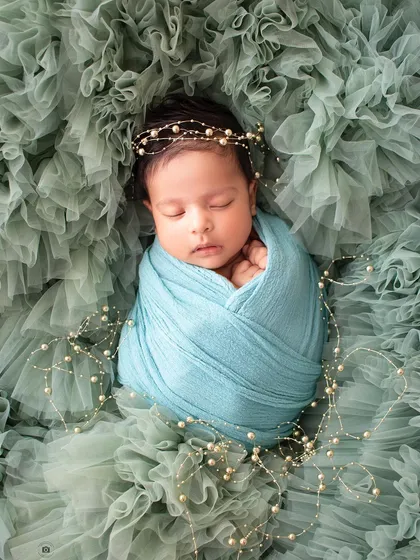 A sleeping newborn is swaddled in turquoise and adorned with a delicate pearl headband, resting on a bed of ruffled green fabric.