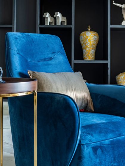 A close-up of the vibrant blue armchair in the Filigree House. The rich velvet texture and the collection of curated decor objects on the shelves behind it highlight the layers of detail in the design.