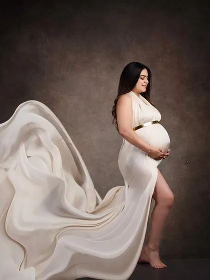 A dramatic fine art portrait featuring a white gown with a flowing train. The fabric is tossed to create a sense of movement against the dark, painterly backdrop.