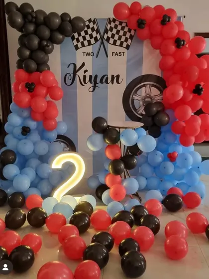 A "Two Fast" race car theme for a second birthday. The decoration includes a custom backdrop and balloon arches in red, blue, and black, with a light-up number '2'.