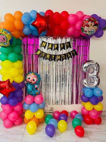 A simple yet colorful Cocomelon theme for a 3rd birthday. This decoration uses a rainbow balloon arch over a shimmer curtain, with character balloons and a large foil number '3'.