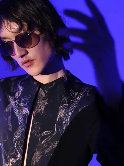 A close-up from a menswear campaign with a cool, moody vibe. The focus is on the printed shirt and sunglasses, with the dramatic blue lighting adding to the overall editorial feel of the shot.