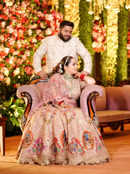 A regal portrait of the couple, with the bride seated and the groom standing behind her, against a beautiful floral backdrop.