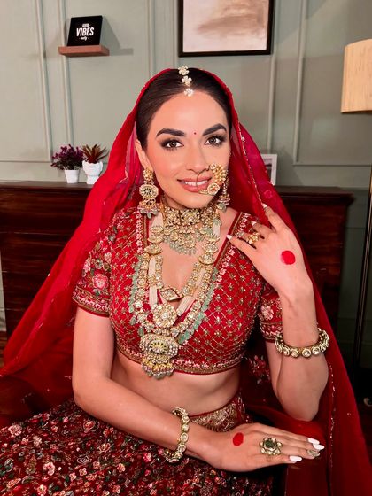 A look of serene confidence. The makeup is designed to complement the rich red lehenga and intricate jewellery, creating a harmonious and regal bridal style.