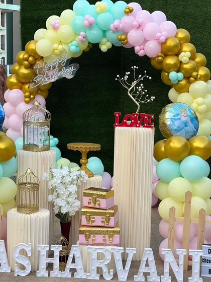A daytime view of the 'Diyasharvani' first birthday setup. You can clearly see the mix of pastel and chrome gold balloons in the arch and the arrangement of pedestals for the cake and other decor.