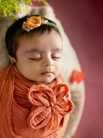 A close-up of a newborn's face, wrapped in a warm orange swaddle. The focus is on their peaceful sleep and the soft, comforting textures.
