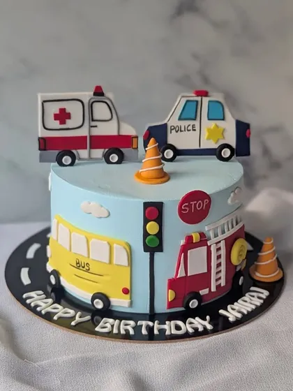 A cute transport-themed cake featuring a police car, ambulance, school bus, and fire truck. It's a colorful and fun design for a young vehicle enthusiast.