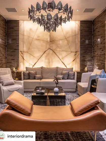 This living room, featured by Interior Drama, showcases an onyx backdrop that adds a significant luxe quotient. The backlit natural stone becomes the focal point, complemented by a sculptural chandelier and a mix of leather and fabric seating.