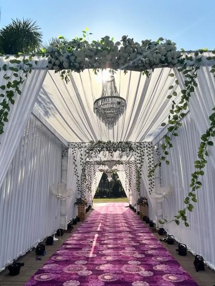 A grand wedding passage for an evening event. The tunnel is created with white drapes, hanging greenery, and chandeliers, leading to the main ceremony area.