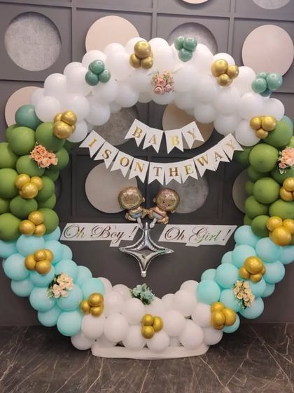 An elegant gender reveal or baby shower theme. The circular balloon arch uses a sophisticated palette of green, teal, and white with gold accents, and a banner that reads 'Baby is on the way'.