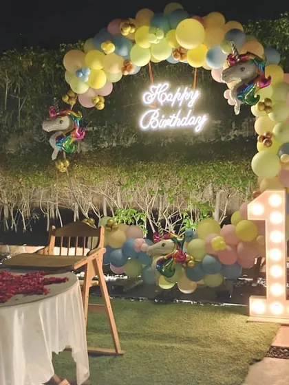A magical outdoor unicorn-themed first birthday party at night. The pastel balloon arch is lit by a neon sign, with a large light-up number '1' adding to the glow.