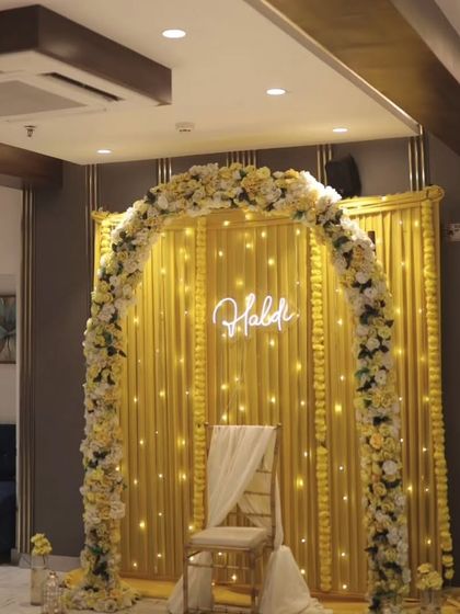 An elegant indoor Haldi setup with a golden fabric backdrop and a floral arch of yellow and white roses. The "Haldi" neon sign adds a modern and personalized touch to the decor.