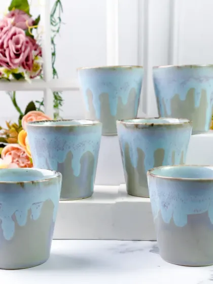 A set of six ceramic tumblers with a beautiful blue drip glaze, arranged on white risers. The floral background adds a touch of softness to the clean, modern presentation of this drinkware.