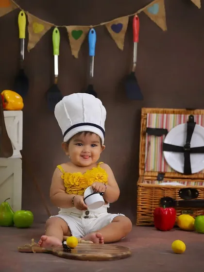 A happy little chef in the making. This sitter session features a kitchen theme, complete with a chef's hat, picnic basket, and colorful toy vegetables for a playful setup.