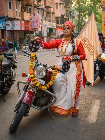 A woman in traditional Maharashtrian attire, including a red and white saree and turban, confidently sits on a decorated motorcycle during the Gudi Padwa festival procession in Girgaon, Mumbai.