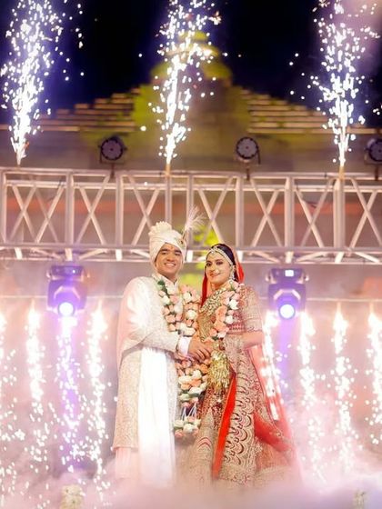 A grand portrait of the couple on stage after their varmala, surrounded by smoke and lights. This is the hero shot that captures the culmination of the ceremony.
