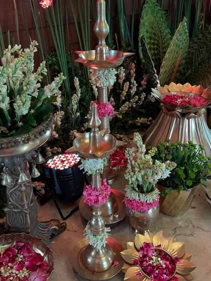 A traditional brass 'diya' stand adorned with fresh flowers, surrounded by other brass decor elements and lush greenery.