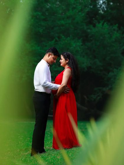 An intimate moment between expectant parents during an outdoor maternity session. The vibrant red dress adds a beautiful pop of color against the lush green background.