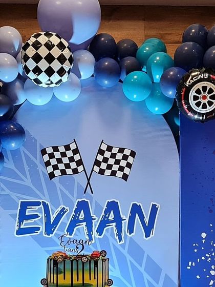 A cool race car-themed birthday party for Evaan. The backdrop features checkered flags and a custom name design, framed by a blue and black balloon garland.